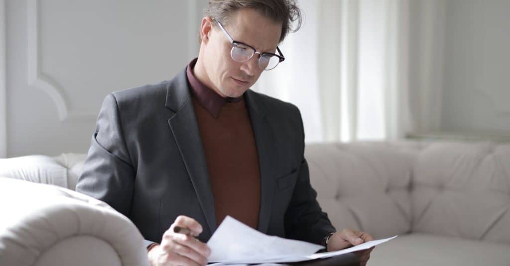 elegant adult man in jacket and glasses looking through documents while sitting on white sofa in lux